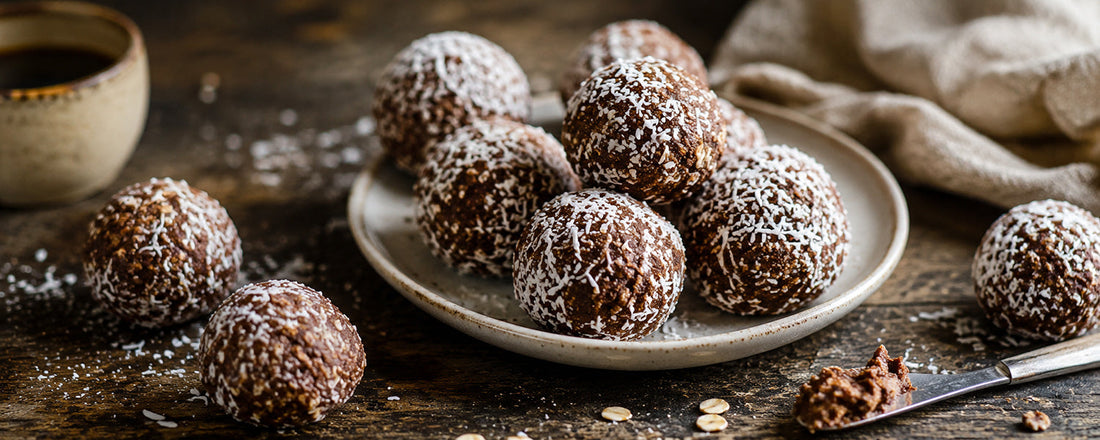 Schwedische Chokladbollar mit Kokosflocken auf einem Keramikteller, serviert auf einem rustikalen Holztisch mit Kaffee und Leinentuch im Hintergrund.