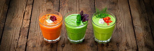 Drei bunte kalte Suppen im Glas – orange, grün und hellgrün – mit frischen Kräutern und Toppings auf einem rustikalen Holztisch.