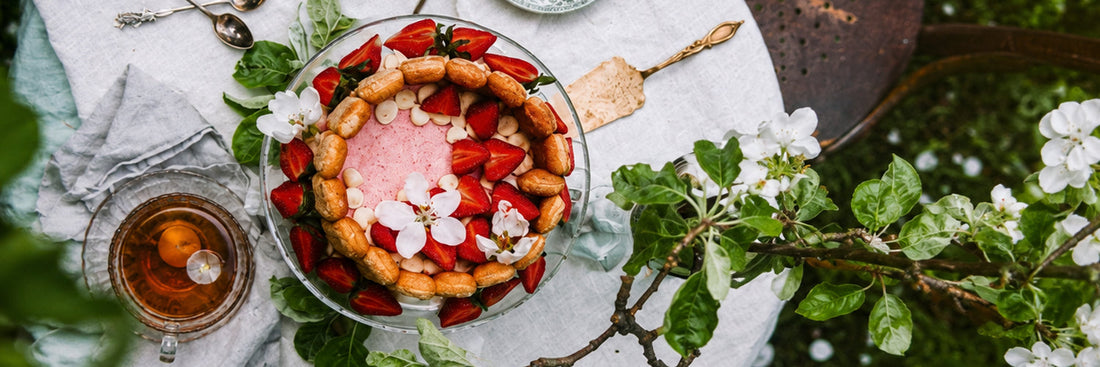 Sommerliche Erdbeertorte mit Blüten auf einem weiß gedeckten Gartentisch, dekoriert mit grün gemusterten Tellern und Tee