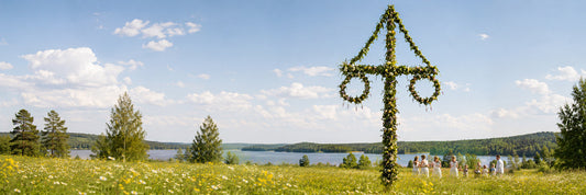 Midsommar-Feier auf einer blühenden Sommerwiese am See mit geschmückter Midsommarstång, schwedischen Flaggen und Menschen in traditioneller Kleidung