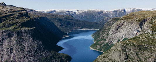 Dramatische Fjordlandschaft in Norwegen: Tiefblauer See zwischen steilen Felswänden und schneebedeckten Bergen im Hintergrund.
