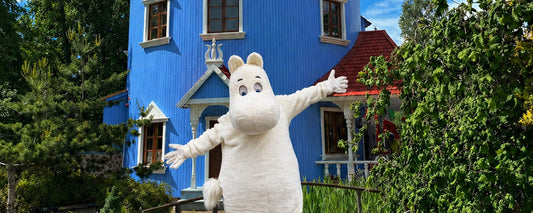 Weiße Mumin-Figur vor blauem Holzhaus in grüner Umgebung