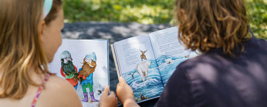 Zwei Kinder sitzen im Grünen auf einer Decke und schauen gemeinsam in farbenfrohe, illustrierte Kinderbücher.